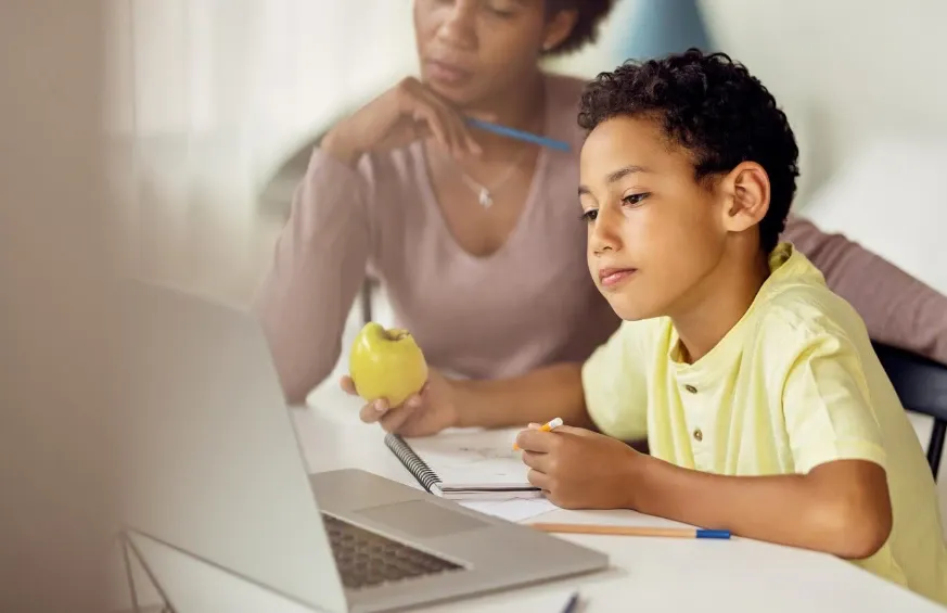 Criança em frente ao computador participa de uma atividade escolar, segurando lápis e observando documentos, enquanto uma professora ao fundo acompanha e ajuda.