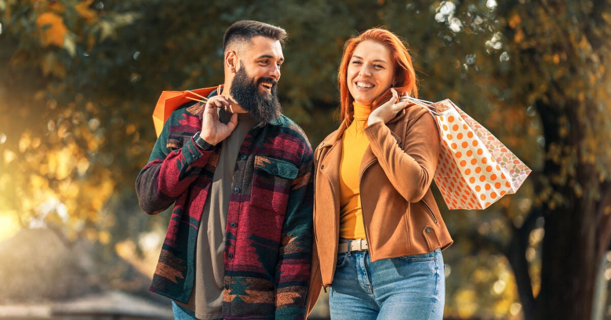 Casal feliz segurando sacolas de compras em um parque outonal, cercado por folhas secas e árvores coloridas.