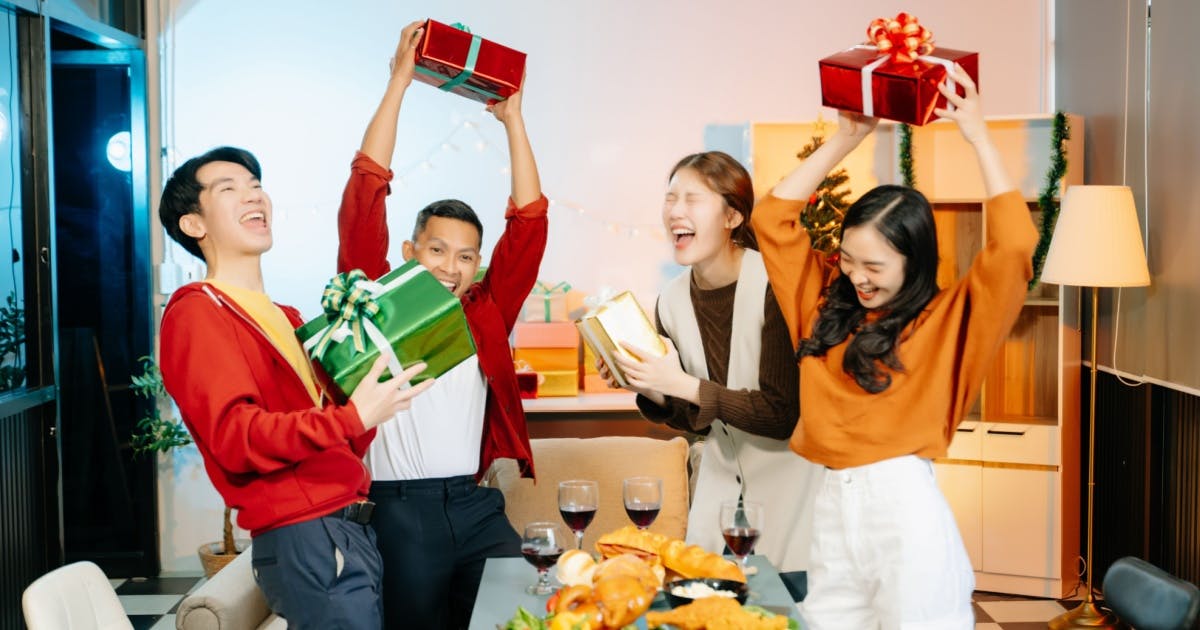 Grupo de amigos de pé sorrindo e comemorando, enquanto erguem seus presentes de Natal, em uma sala, em torno de uma mesa.