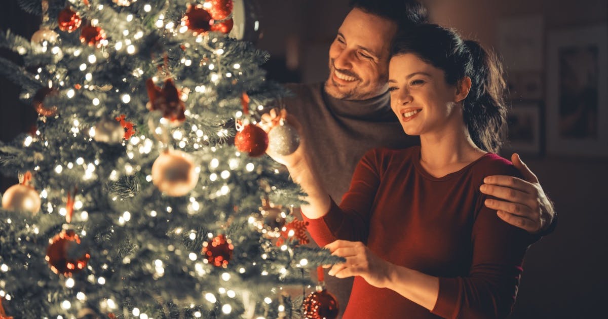 Casal de pé sorrindo, decorando uma árvore de Natal, enquanto o homem passa o braço por cima do ombro da mulher.