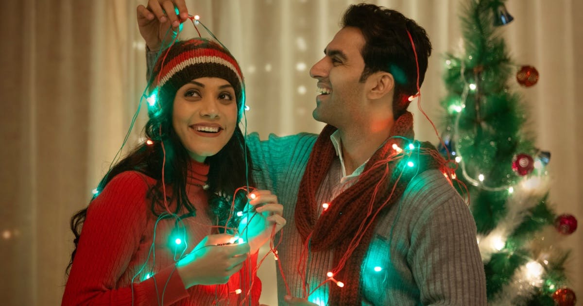 Casal sorrindo, enrolados em pisca-piscas natalinos, enquanto decoram uma casa com uma árvore de Natal ao fundo.