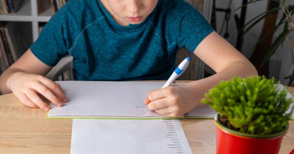 Menino escrevendo em um caderno com uma caneta, praticando o seu vocabulário.