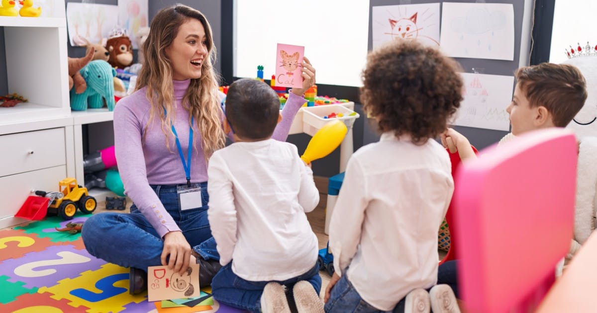 Mulher ensina vocabulário para crianças em uma sala de aula, rodeada de mesas e quadros, promovendo aprendizado e interação.