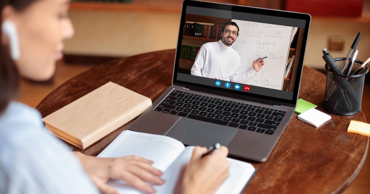 Mulher sentada à mesa com um laptop e, na tela, um homem explicando o conteúdo do curso de inglês online.