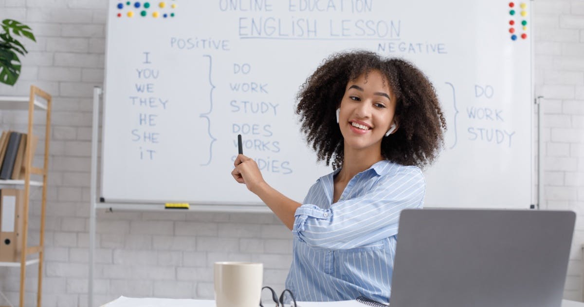 Mulher sorrindo diante de um quadro branco, enquanto dá uma aula em um curso de inglês online.