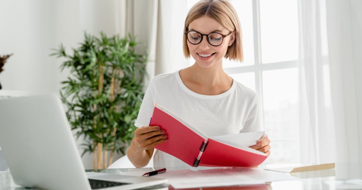 Mulher de óculos segurando um caderno e olhando para o laptop, durante uma aula de um curso de inglês online.