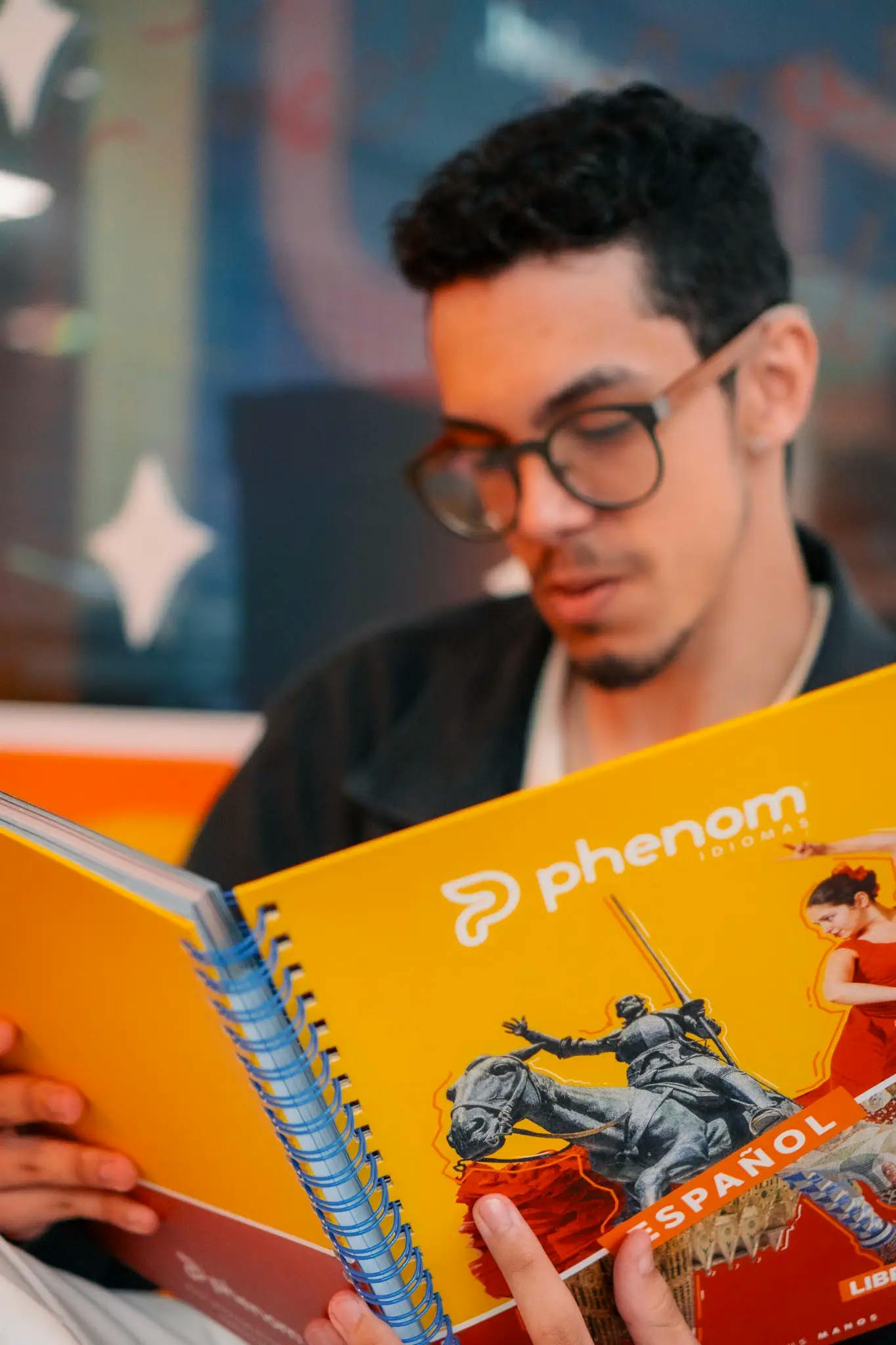 Homem jovem com óculos lendo um material didático amarelo da Phenom Idiomas, com destaque para o texto 'Espanhol' e imagens temáticas relacionadas à cultura espanhola na capa.