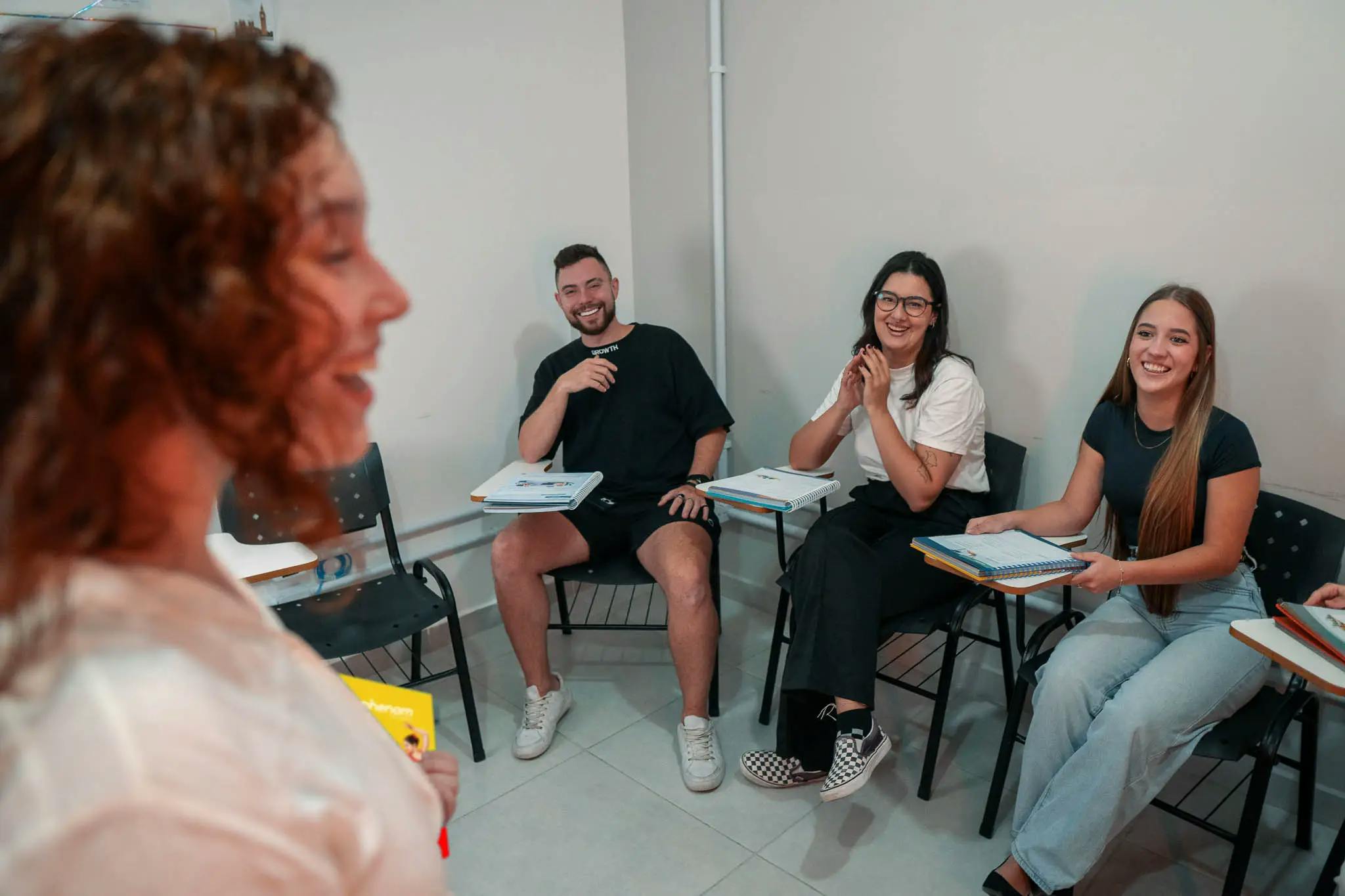 Professora interagindo com alunos sorridentes durante uma aula em grupo na Phenom Idiomas, com materiais didáticos visíveis sobre as mesas.