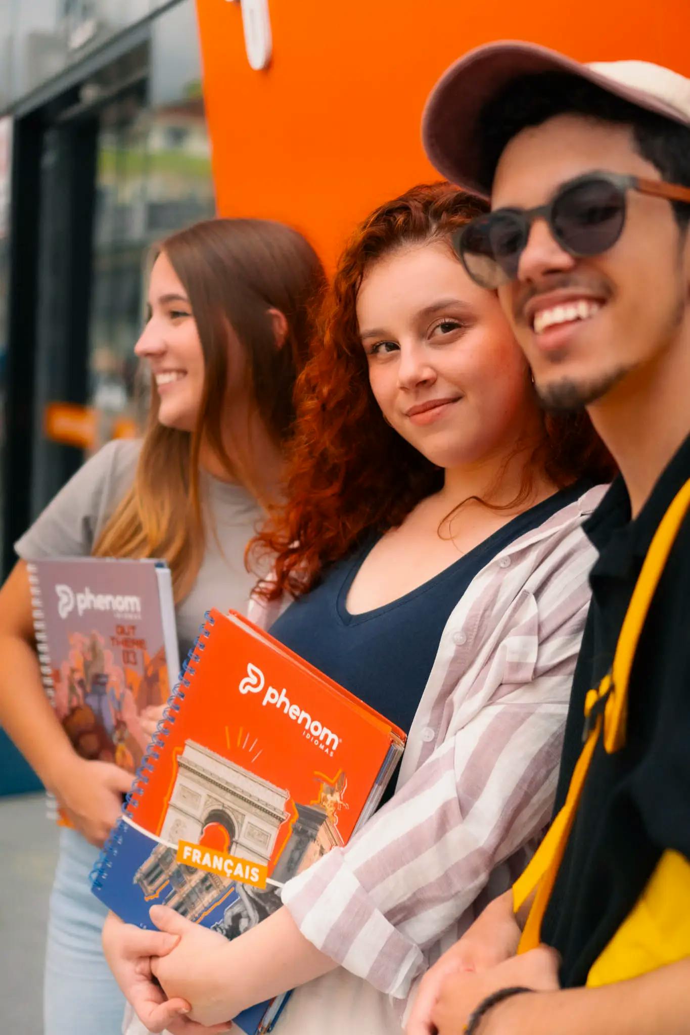 Três jovens sorrindo, segurando materiais didáticos da Phenom Idiomas, incluindo um livro de francês com a imagem do Arco do Triunfo, em frente a um fundo laranja vibrante.