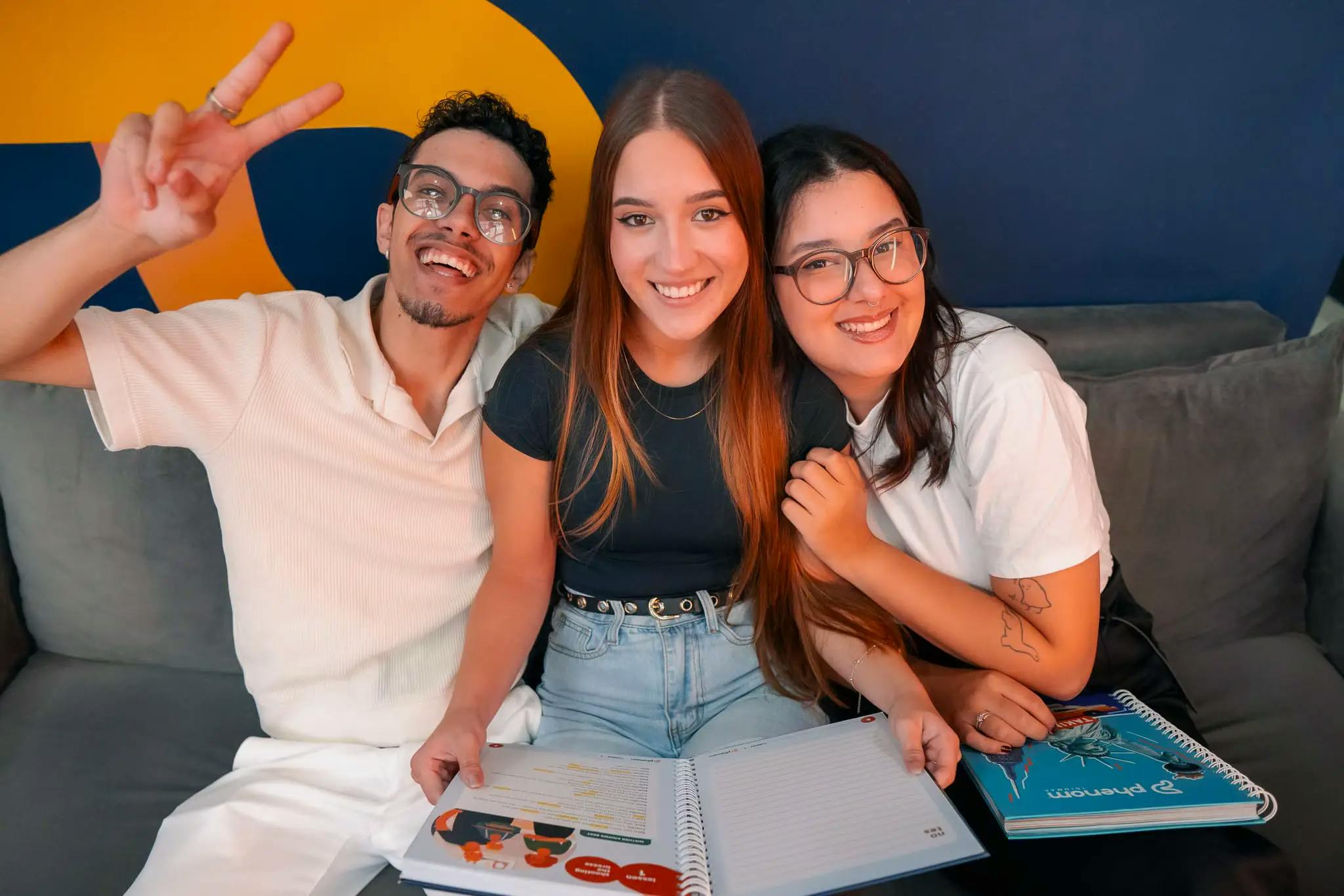 Três jovens sorrindo e sentados juntos em um sofá, segurando materiais didáticos da Phenom Idiomas, com um fundo azul e amarelo vibrante.