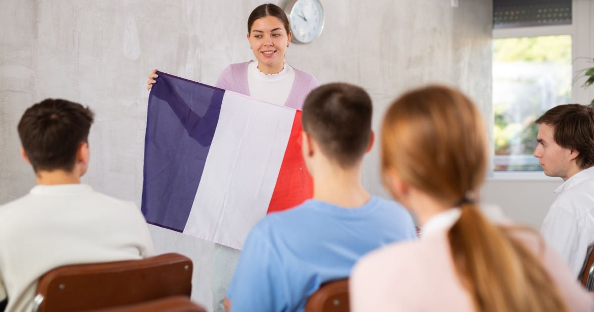 Mulher segurando uma bandeira francesa na frente de um grupo de alunos, durante uma aula de francês.