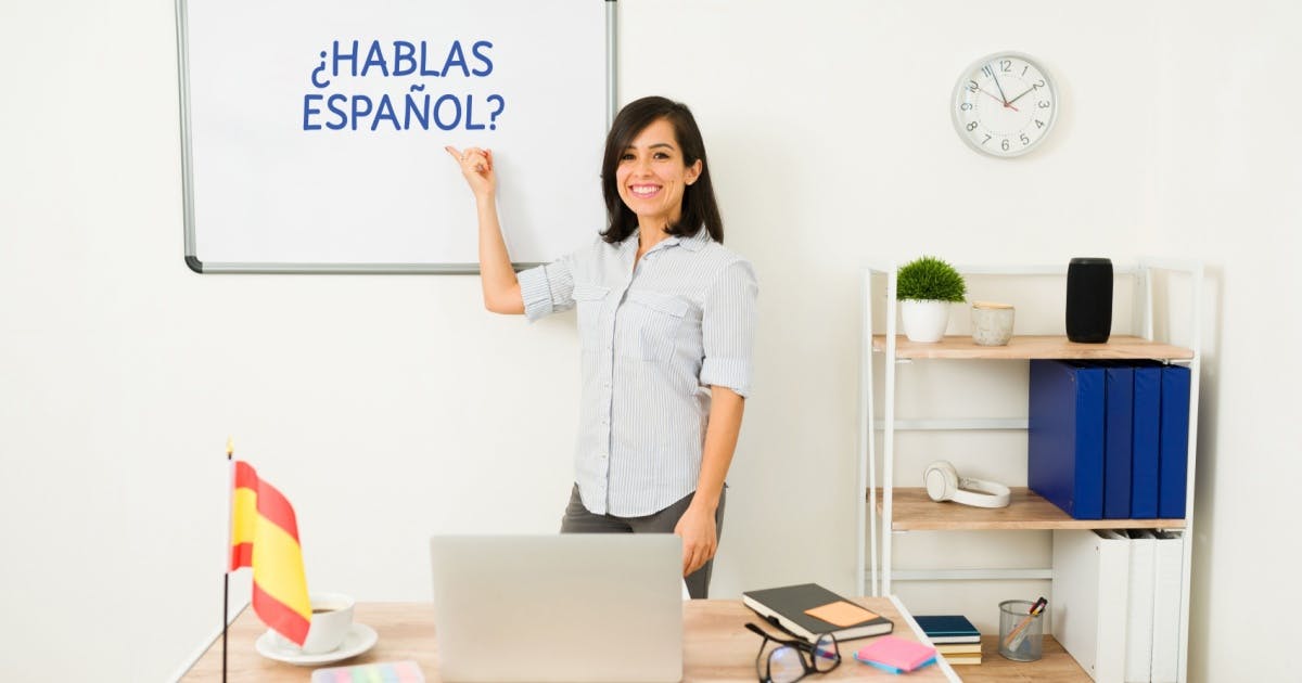 Mulher anotando em um quadro branco, dando aula de espanhol, com um ambiente de aprendizado ao fundo.