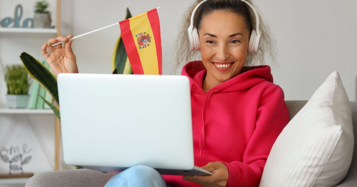 Mulher sentada, segurando uma bandeira da Espanha e um notebook para uma aula de espanhol online.