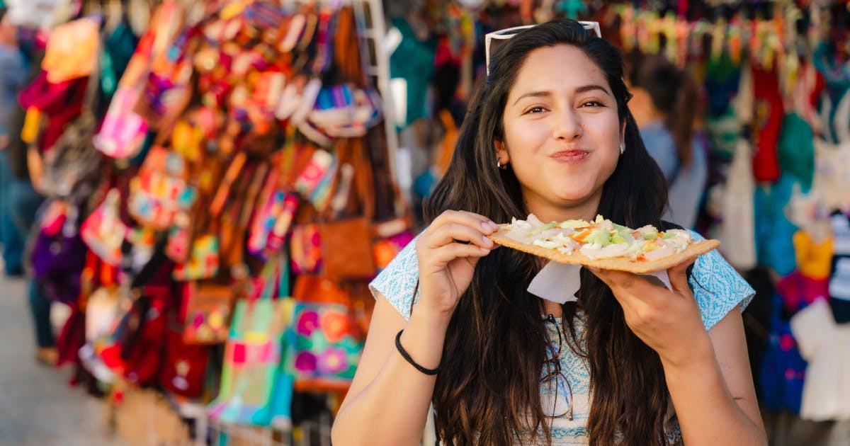 Uma mulher saboreando uma comida típica em frente a um mercado movimentado.