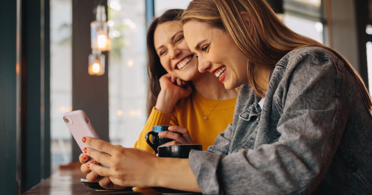 Duas mulheres sentadas à mesa, concentradas em seus celulares, compartilhando um momento de descontração.