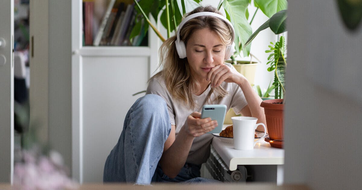 Uma mulher sentada no sofá, usando fones de ouvido e segurando uma xícara de café.