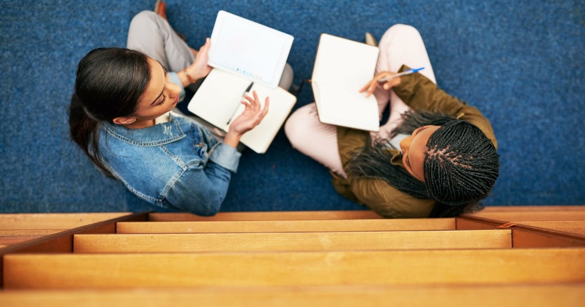 Duas alunas com livros nas mãos e conversando. O ângulo da foto é com a vista de cima.