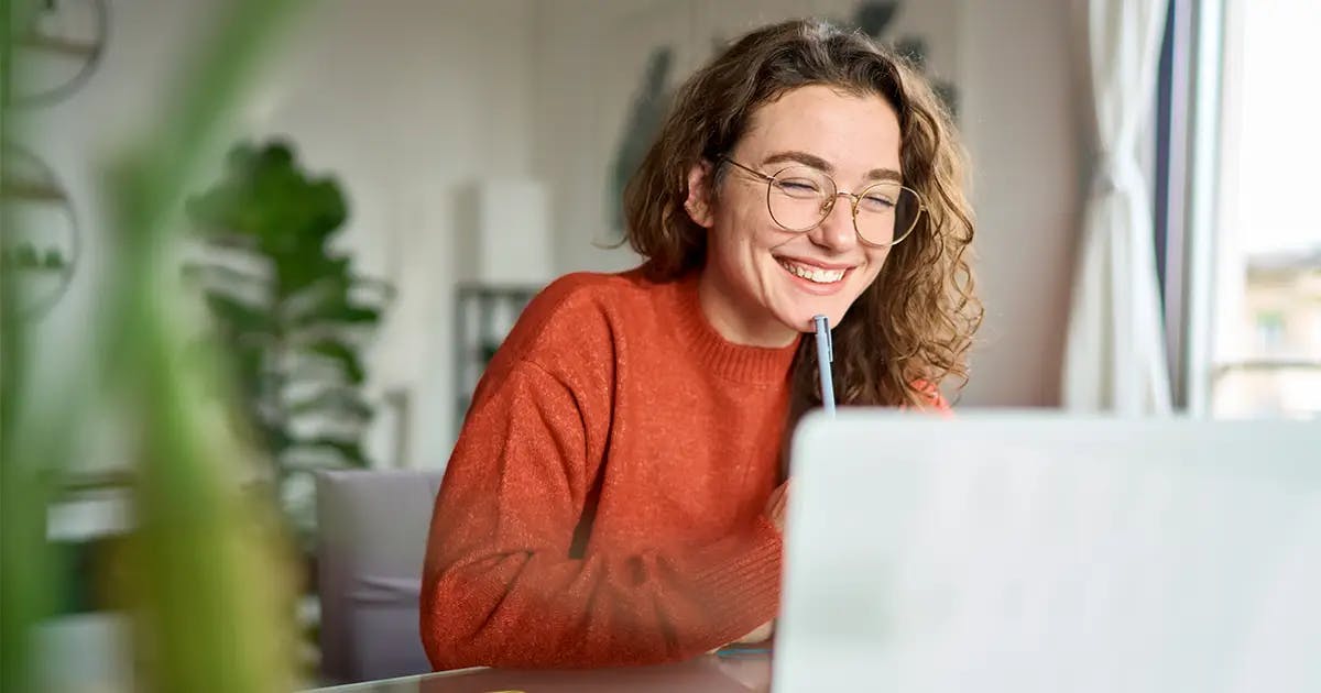 Mulher jovem com óculos e suéter laranja sorrindo enquanto estuda em um notebook, segurando uma caneta, em um ambiente aconchegante com plantas ao fundo.