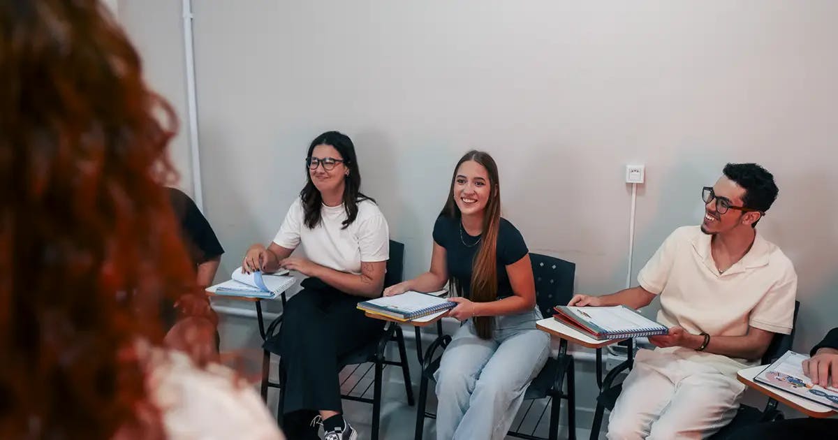 Grupo de jovens estudantes sorrindo e participando de uma aula interativa, segurando materiais didáticos da Phenom Idiomas, com a professora parcialmente visível em primeiro plano.