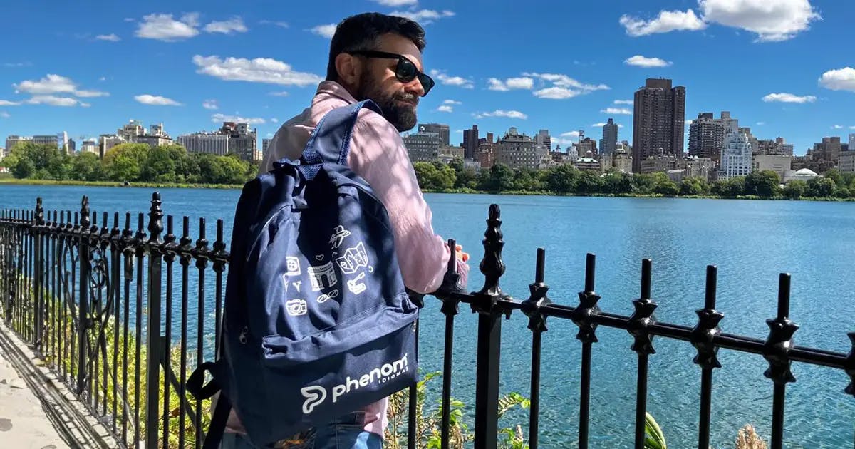 Homem com óculos escuros e mochila da Phenom Idiomas encostado em uma grade, admirando a paisagem urbana com um lago e prédios ao fundo em um dia ensolarado.