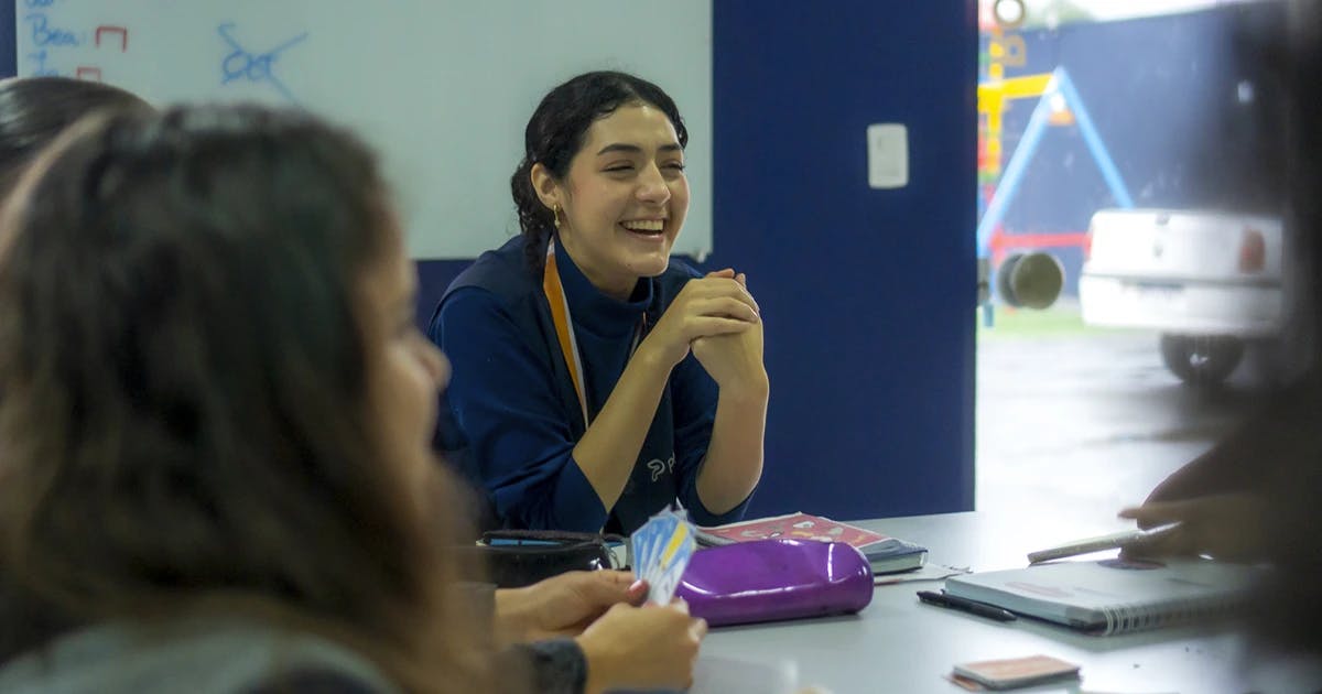 Aluna sorridente participando de uma atividade em grupo na Phenom Idiomas, sentada à mesa com materiais de estudo e cartas educativas, em um ambiente descontraído e interativo.