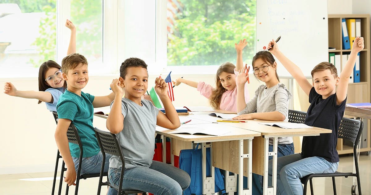 Crianças sorridentes em uma sala de aula moderna, sentadas ao redor de uma mesa, levantando as mãos com entusiasmo e segurando pequenos materiais de estudo, com uma bandeira dos EUA ao fundo.