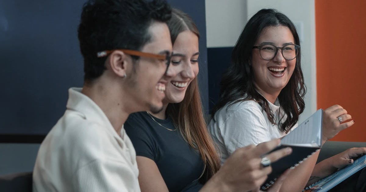 Três jovens sorrindo enquanto estudam juntos, segurando materiais didáticos da Phenom Idiomas, em um ambiente descontraído e colaborativo.