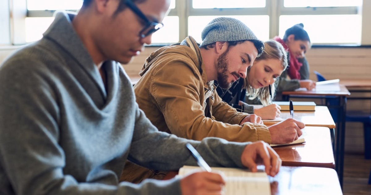 Grupo de estudantes sentados em carteiras, concentrados e escrevendo em folhas de papel, enquanto faz prova de inglês.