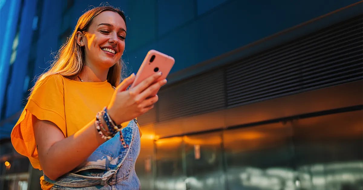 Mulher feliz vestindo camiseta amarela e macacão jeans verificando seu celular ao ar livre durante iluminação dourada