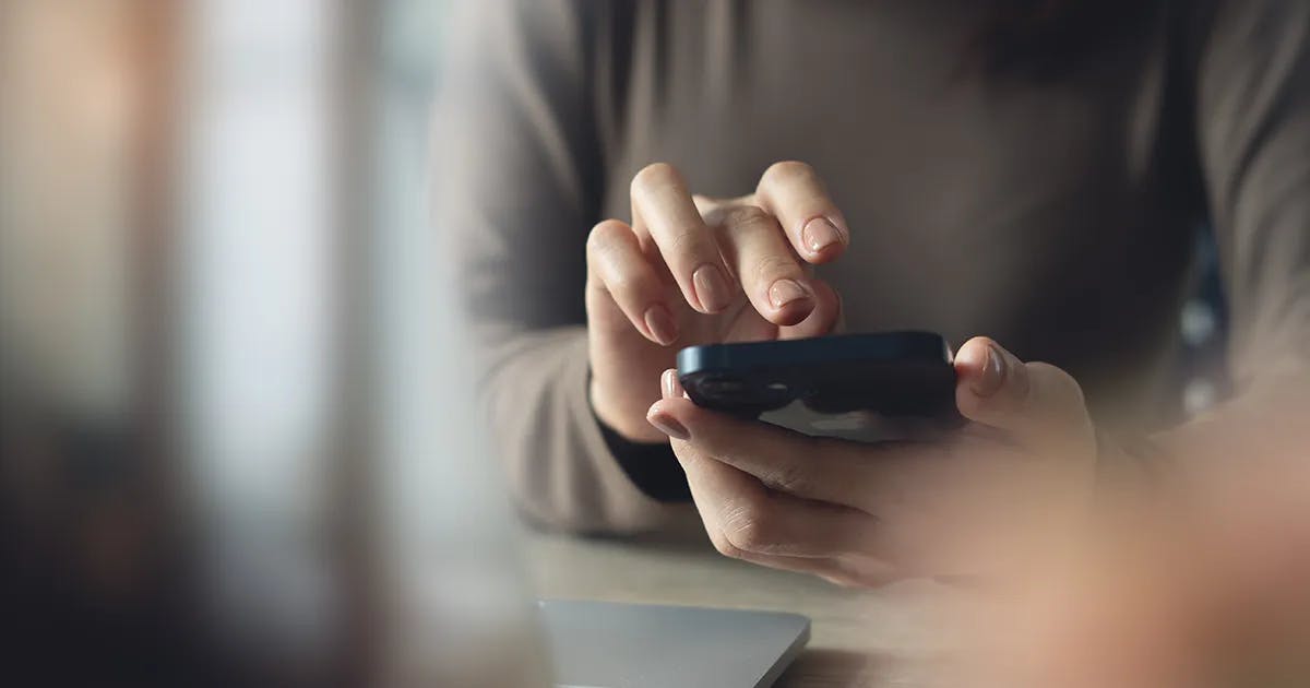 Close-up de mãos segurando e digitando em um smartphone preto sobre uma mesa, em ambiente com iluminação suave