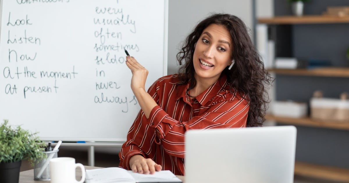 Professora sorrindo, dando aula online, enquanto olha para a tela do notebook e aponta para as informações na lousa branca.