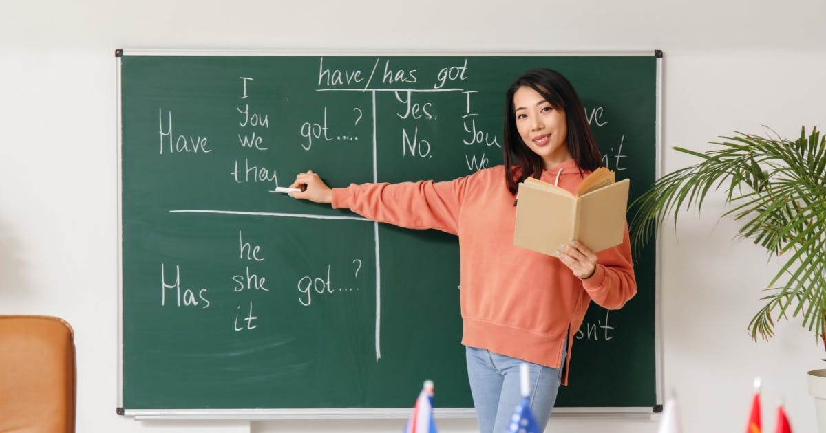 Professora de pé na frente, segurando um livro aberto e apontando um giz para uma explicação sobre conversação em inglês.