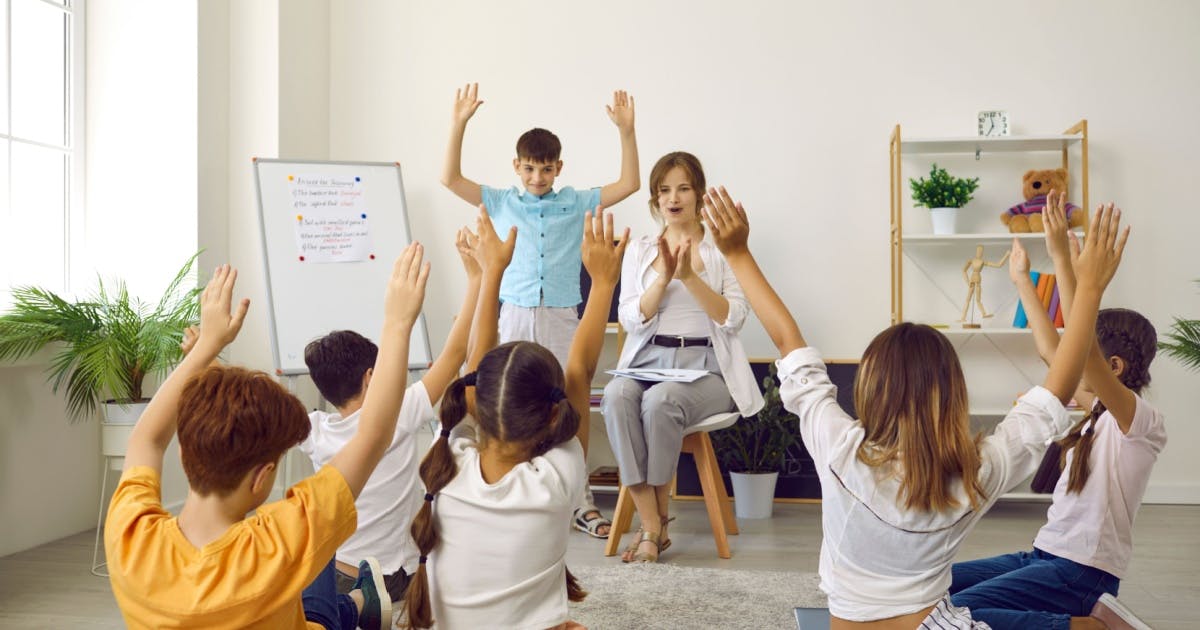 Professora sentada, dando aula de inglês para uma turma de crianças que estão com os braços levantados participando da aula.