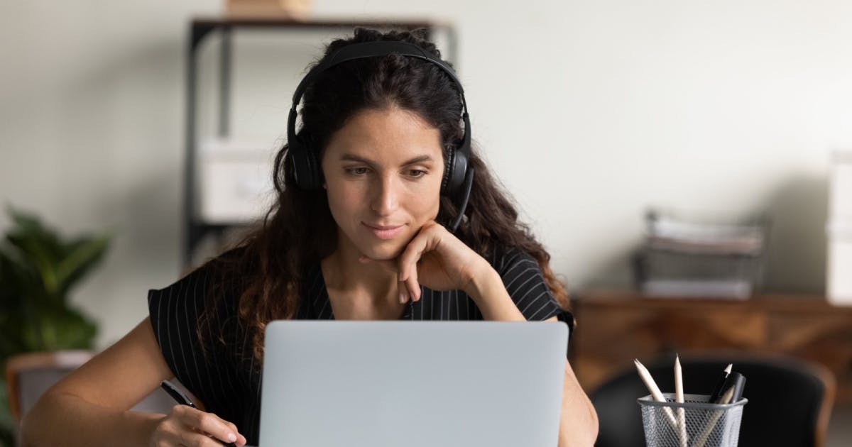 Mulher de fones de ouvido, sentada, observando um notebook aberto, enquanto estuda ouvindo músicas em inglês.