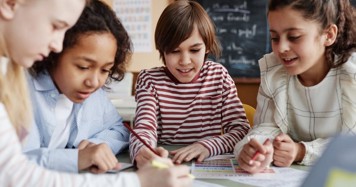 Crianças alunas de uma escola de inglês estudando juntas, reunidas em volta de uma mesa, com um quadro negro ao fundo.