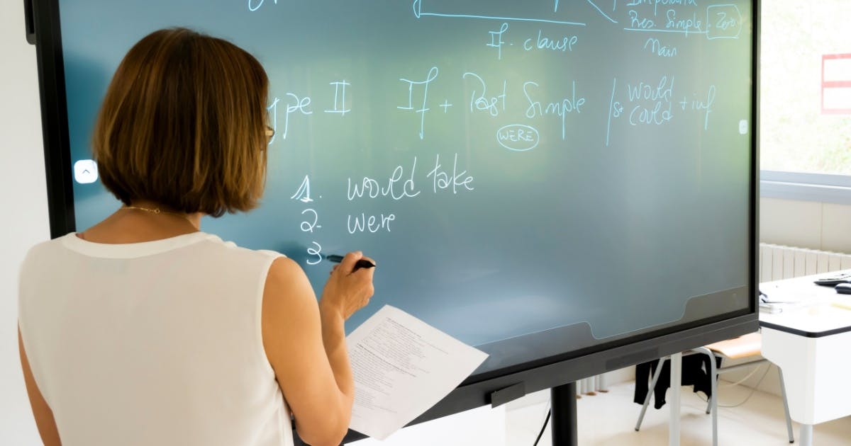 Professora em uma escola de inglês, segurando uma folha e apontando uma caneta para uma tela para dar uma explicação.