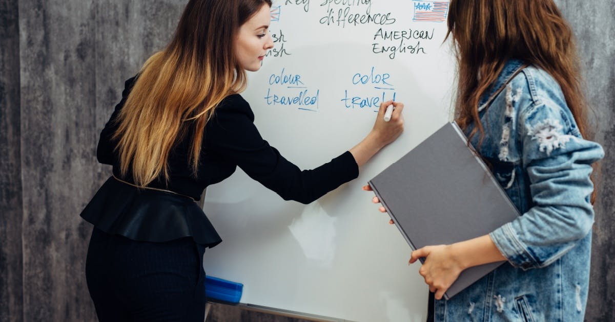 Professora e aluna de pé, enquanto a professora ensina a aluna a falar inglês, dando explicação escrevendo na lousa.