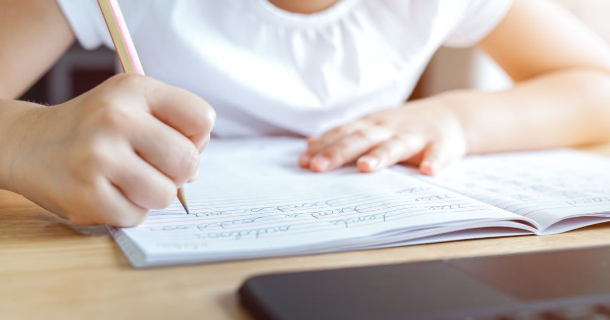 Criança de camiseta branca sentada, aprendendo a escrever em inglês em um caderno apoiado em uma mesa de madeira.