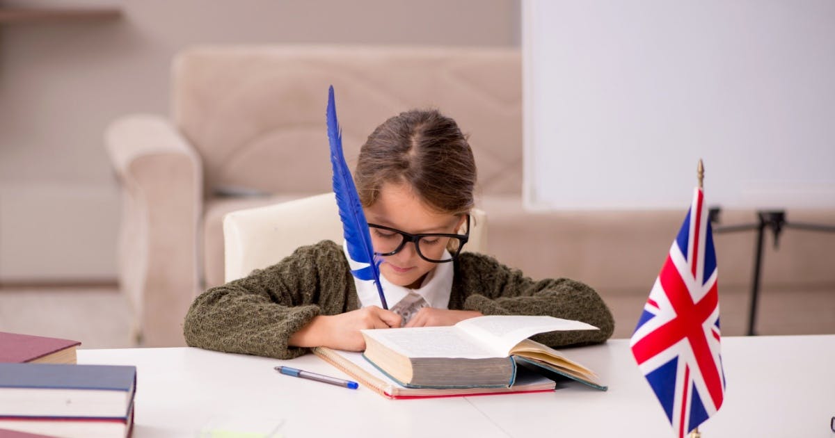 Criança aprendendo a escrever em inglês em um caderno apoiado em uma mesa com alguns livros e uma bandeira da Grã-Bretanha.