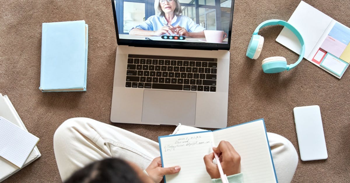 Aluna sentada na frente de um notebook aberto, tendo aula online e anotando explicação sobre gírias em inglês em um caderno.