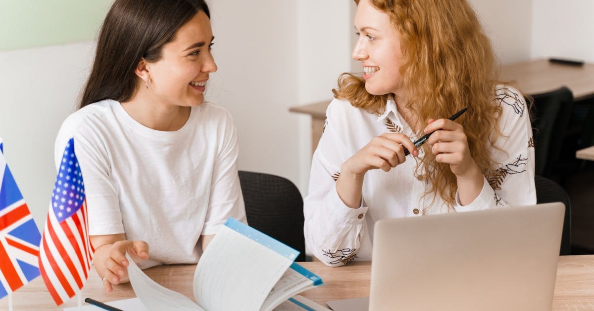 Professora e aluna sentadas na sala de aula, conversando enquanto a professora avalia a pronúncia em inglês da aluna.