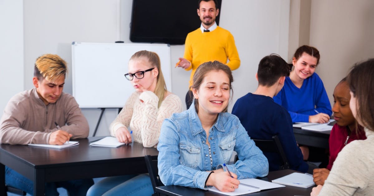 Grupo de alunos sentados, conversando enquanto o professor está de pé aplicando seu método de estudo e os observando.