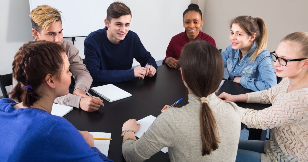 Grupo de alunos sentados, conversando reunidos em volta de uma mesa com cadernos para fazer anotações do método de estudo.