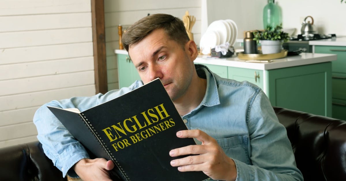 Homem sentado em um sofá, enquanto lê um livro em inglês para iniciantes com a frase "English for beginners" na capa.