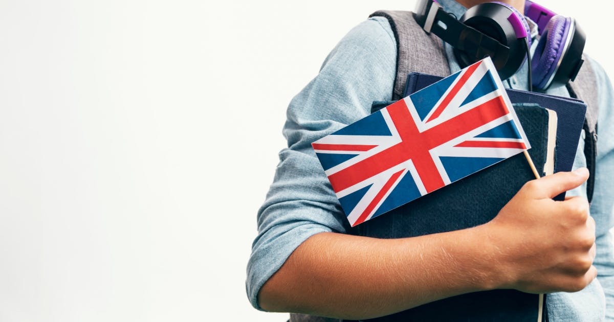 Homem de pé na frente de um fundo branco, com um headphone no pescoço, enquanto segura livros para estudar inglês britânico.