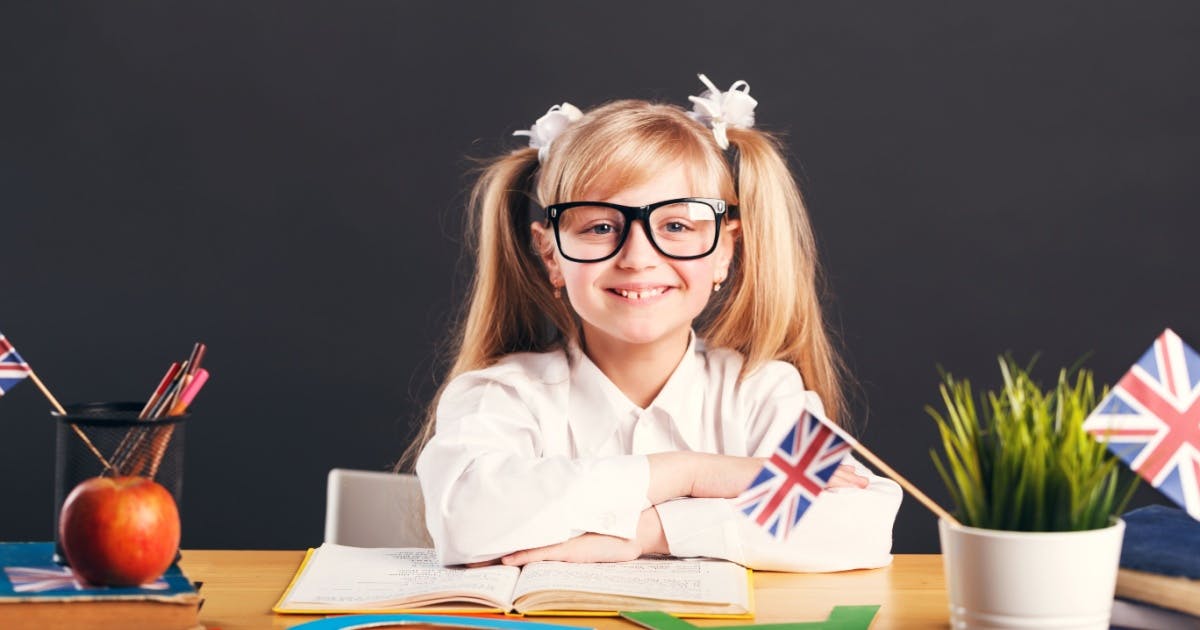 Menina de óculos, estudante de inglês britânico, em uma sala de aula, sentada e apoiada em uma mesa com alguns objetos.