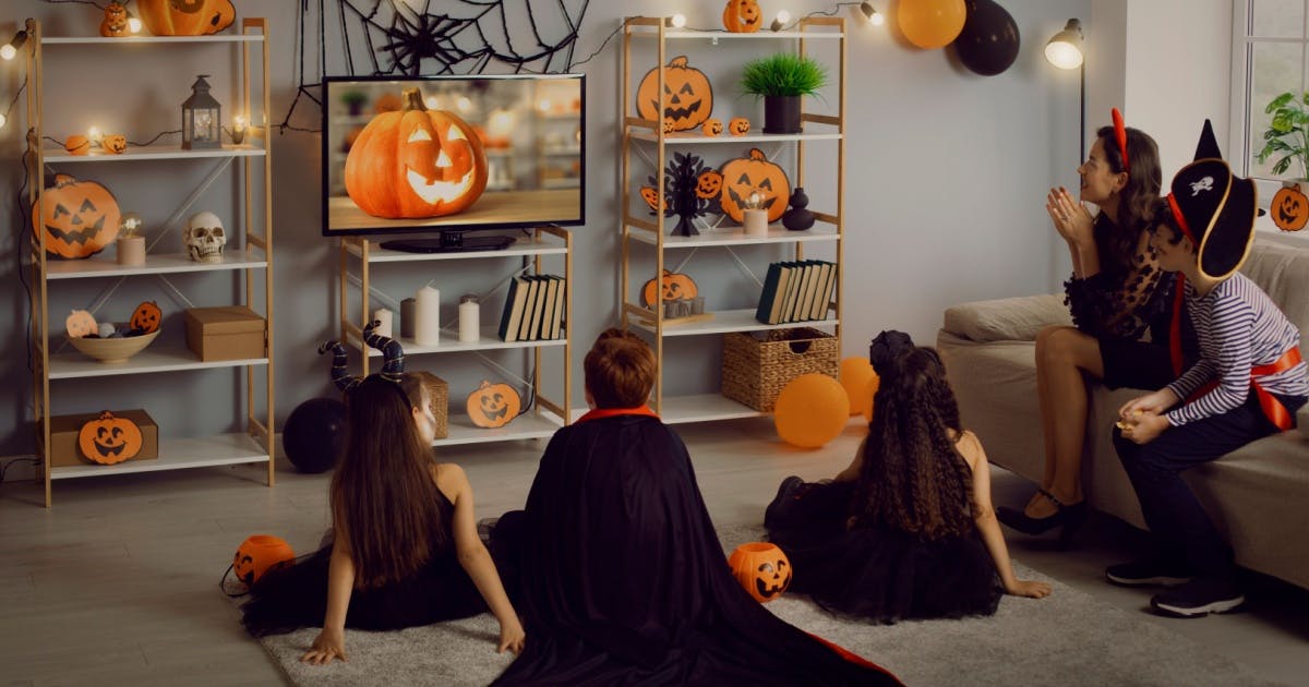 Uma mulher e quatro crianças sentadas e fantasiadas, assistindo a um filme de terror no Halloween em uma sala decorada.