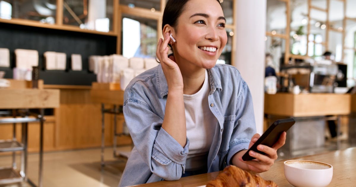 Mulher sentada e sorrindo, enquanto segura o smartphone e escuta uma lição de um aplicativo para aprender inglês.