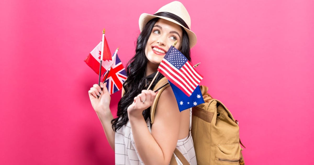 Mulher de pé, sorrindo com uma mochila nas costas e um chapéu, enquanto segura bandeiras de países que falam inglês.