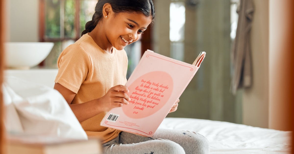 Menia sentada no sofá, lendo um livro infantil em inglês, tentando aprender inglês sozinha.